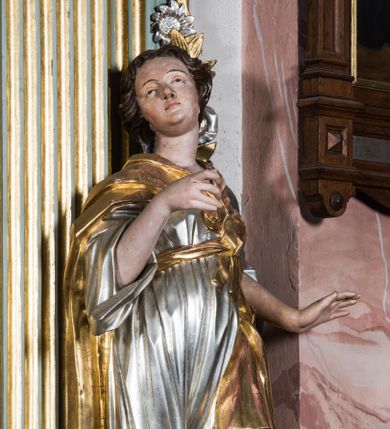 Zdjęcie nr 1: Pełnopostaciowa figura ukazuje stojącą frontalnie świętą niewiastę w kontrapoście. Święta z odchyloną nieznacznie do tyłu głową prawą ręką unosi na wysokości piersi trzymając w niej niezachowany przedmiot, lewą zaś lekko odsuwa od siebie. Kobieta ma pociągłą twarz o pucułowatych, rumianych policzkach, wyrazisty nos i usta. Jej głowę otaczają brązowe włosy spływające na ramiona. Ubrana jest w srebrzoną tunikę przewiązaną na piersi szarfą oraz narzucony na ramiona złoty płaszcz osuwający się z tyłu postaci. Spod tuniki widać fragment lewej stopy obutej w sandał.