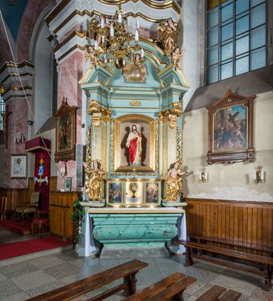 Zdjęcie nr 2: Przyścienny, wczesnoklasycystyczny ołtarz uzyskał formę architektonicznej, jednokondygnacyjnej nastawy ze zwieńczeniem. Sarkofagowy stipes ołtarzowy podtrzymuje drewnianą mensy, na której ustawiono prostopadłościenne tabernakulum w formie skrzynki. Złocone drzwiczki ozdobiono płaskorzeźbionym przedstawieniem kielicha ustawionego na obłokach z hostią otoczoną promieniami. Tabernakulum flankują dwie wypukło wklęsłe płyciny zawierające obraz ze scena pokłonu Trzech Króli oraz Zmartwychwstanie Chrystusa. Powyżej wzniesiono retabulum artykułowane po bokach kanelowanymi pilastrami z kompozytowymi głowicami, podtrzymującymi wyłamane po zewnętrznych belkowanie, dodatkowo wsparte na ustawionych w narożach konsolach. Konsole ozdobiono ornament z motywem  stylizowanej muszli. Na belkowaniu wsparto przerwany przyczółek, na bokach którego siedzą dwa uskrzydlone anioły. Zwieńczenie utworzono z lezącego prostokąta o wklęsłych bokach zwieńczonego profilowanym gzymsem ujętym w nadwieszony łuk odcinkowy. Na jego szczycie po bokach ustawiono dwa stojące uskrzydlone anioły. W polu zwieńczenia znalazł się płaskorzeźbiony chrystogram "IHS" w promienistej glorii. Powyżej umieszczono parę uskrzydlonych główek anielskich. Z kolei w polu kondygnacji między pilastrami usytuowano półokrągłą niszę z figurą Najświętszego Serca Pana Jezusa. Na tle pilastrów dostawione zostały posągi św. Barbary i św. Marii Magdaleny. Struktura została pomalowana w kolorze jasnego seledynu, ornamenty i profile wyzłocono, a rzeźby otrzymały naturalistyczną polichromię.