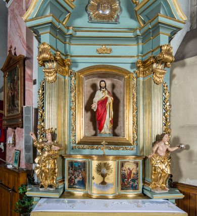 Zdjęcie nr 3: Przyścienny, wczesnoklasycystyczny ołtarz uzyskał formę architektonicznej, jednokondygnacyjnej nastawy ze zwieńczeniem. Sarkofagowy stipes ołtarzowy podtrzymuje drewnianą mensy, na której ustawiono prostopadłościenne tabernakulum w formie skrzynki. Złocone drzwiczki ozdobiono płaskorzeźbionym przedstawieniem kielicha ustawionego na obłokach z hostią otoczoną promieniami. Tabernakulum flankują dwie wypukło wklęsłe płyciny zawierające obraz ze scena pokłonu Trzech Króli oraz Zmartwychwstanie Chrystusa. Powyżej wzniesiono retabulum artykułowane po bokach kanelowanymi pilastrami z kompozytowymi głowicami, podtrzymującymi wyłamane po zewnętrznych belkowanie, dodatkowo wsparte na ustawionych w narożach konsolach. Konsole ozdobiono ornament z motywem  stylizowanej muszli. Na belkowaniu wsparto przerwany przyczółek, na bokach którego siedzą dwa uskrzydlone anioły. Zwieńczenie utworzono z lezącego prostokąta o wklęsłych bokach zwieńczonego profilowanym gzymsem ujętym w nadwieszony łuk odcinkowy. Na jego szczycie po bokach ustawiono dwa stojące uskrzydlone anioły. W polu zwieńczenia znalazł się płaskorzeźbiony chrystogram "IHS" w promienistej glorii. Powyżej umieszczono parę uskrzydlonych główek anielskich. Z kolei w polu kondygnacji między pilastrami usytuowano półokrągłą niszę z figurą Najświętszego Serca Pana Jezusa. Na tle pilastrów dostawione zostały posągi św. Barbary i św. Marii Magdaleny. Struktura została pomalowana w kolorze jasnego seledynu, ornamenty i profile wyzłocono, a rzeźby otrzymały naturalistyczną polichromię.