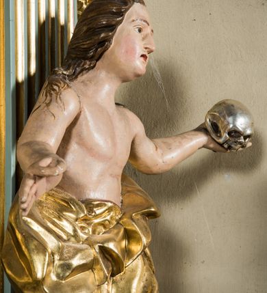 Zdjęcie nr 3: Pełnopostaciowa figura ukazuje stojącą frontalnie świętą Marię Magdalenę w kontrapoście. Święta rozkłada ręce, trzymając w lewej dłoni srebrzoną czaszkę. Kobieta ma pociągłą twarz o pucułowatych, rumianych policzkach, wyrazisty nos i usta oraz szeroko otwarte oczy ze spojrzeniem skierowanym na widza. Jej głowę otaczają długie brązowe włosy spływające na ramiona. Ubrana jest w złoconą, mocno drapowaną suknie ściągniętą do pasa, odsłaniającą jej nagi tułów i ręce. Spod szaty widać końce bosych stóp.