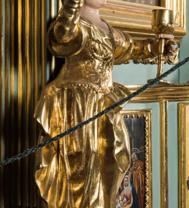 Zdjęcie nr 2: Pełnopostaciowa figura ukazuje stojącą frontalnie świętą Barbarę w kontrapoście. Święta w uniesionej na wysokości piersi lewej ręce trzyma złocony kielich, natomiast prawą ma uniesioną ku górze na wysokość głowy. Kobieta ma pociągłą twarz o pucułowatych, rumianych policzkach, wyrazisty nos i usta oraz szeroko otwarte oczy ze spojrzeniem skierowanym na widza. Jej głowę otaczają długie brązowe włosy spływające na ramiona. Na głowie ma złoconą koronę o floralnych sterczynach. Ubrana jest w złocony, taliowany, wzorzysty gorset oraz mocno drapowaną spódnicę odsłaniającą czubki butów.