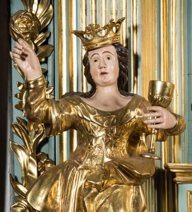 Zdjęcie nr 1: Pełnopostaciowa figura ukazuje stojącą frontalnie świętą Barbarę w kontrapoście. Święta w uniesionej na wysokości piersi lewej ręce trzyma złocony kielich, natomiast prawą ma uniesioną ku górze na wysokość głowy. Kobieta ma pociągłą twarz o pucułowatych, rumianych policzkach, wyrazisty nos i usta oraz szeroko otwarte oczy ze spojrzeniem skierowanym na widza. Jej głowę otaczają długie brązowe włosy spływające na ramiona. Na głowie ma złoconą koronę o floralnych sterczynach. Ubrana jest w złocony, taliowany, wzorzysty gorset oraz mocno drapowaną spódnicę odsłaniającą czubki butów.