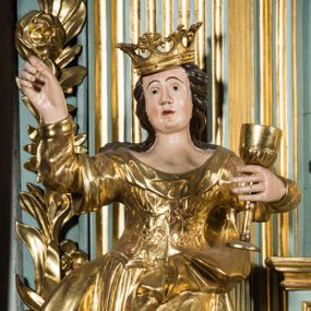 Zdjęcie nr 1: Pełnopostaciowa figura ukazuje stojącą frontalnie świętą Barbarę w kontrapoście. Święta w uniesionej na wysokości piersi lewej ręce trzyma złocony kielich, natomiast prawą ma uniesioną ku górze na wysokość głowy. Kobieta ma pociągłą twarz o pucułowatych, rumianych policzkach, wyrazisty nos i usta oraz szeroko otwarte oczy ze spojrzeniem skierowanym na widza. Jej głowę otaczają długie brązowe włosy spływające na ramiona. Na głowie ma złoconą koronę o floralnych sterczynach. Ubrana jest w złocony, taliowany, wzorzysty gorset oraz mocno drapowaną spódnicę odsłaniającą czubki butów.