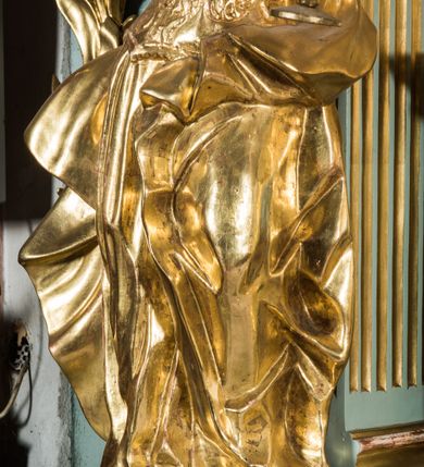 Zdjęcie nr 5: Pełnopostaciowa figura ukazuje stojącą frontalnie świętą Barbarę w kontrapoście. Święta w uniesionej na wysokości piersi lewej ręce trzyma złocony kielich, natomiast prawą ma uniesioną ku górze na wysokość głowy. Kobieta ma pociągłą twarz o pucułowatych, rumianych policzkach, wyrazisty nos i usta oraz szeroko otwarte oczy ze spojrzeniem skierowanym na widza. Jej głowę otaczają długie brązowe włosy spływające na ramiona. Na głowie ma złoconą koronę o floralnych sterczynach. Ubrana jest w złocony, taliowany, wzorzysty gorset oraz mocno drapowaną spódnicę odsłaniającą czubki butów.