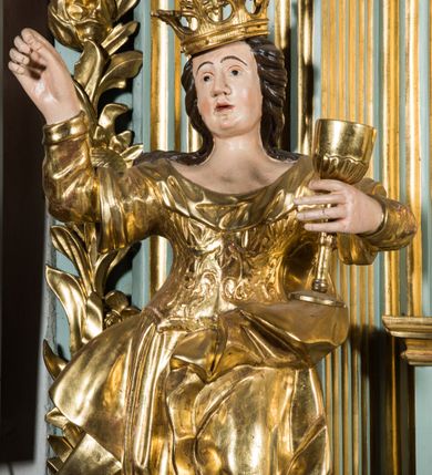 Zdjęcie nr 4: Pełnopostaciowa figura ukazuje stojącą frontalnie świętą Barbarę w kontrapoście. Święta w uniesionej na wysokości piersi lewej ręce trzyma złocony kielich, natomiast prawą ma uniesioną ku górze na wysokość głowy. Kobieta ma pociągłą twarz o pucułowatych, rumianych policzkach, wyrazisty nos i usta oraz szeroko otwarte oczy ze spojrzeniem skierowanym na widza. Jej głowę otaczają długie brązowe włosy spływające na ramiona. Na głowie ma złoconą koronę o floralnych sterczynach. Ubrana jest w złocony, taliowany, wzorzysty gorset oraz mocno drapowaną spódnicę odsłaniającą czubki butów.