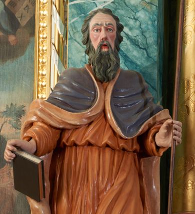 Zdjęcie nr 1: Rzeźba przedstawiająca proroka Eliasza, ścięta z tyłu, drążona. Prorok ustawiony na niskim cokole w formie posrebrzonego obłoku. Ukazany jest frontalnie w delikatnym kontrapoście z lewą nogą ugiętą w kolanie i z rękami ugiętymi w łokciach. W prawej dłoni trzyma zamkniętą księgę, a w lewej laskę. Twarz podłużna z długim nosem, otwartymi ustami i zmarszczonymi łukami brwiowymi; okolona długą, ciemną brodą i średniej długości włosami, zasłaniającymi uszy, silnie podwiniętymi do tyłu. Ubrany jest w długą, jasnobrązową suknię, przewiązaną w talii, obficie drapowaną oraz ciemnobrązowy płaszcz z jasnobrązową podszewką, spięty na piersiach i zarzucony na przedramionach do tyłu; na stopach ma założone pełne, brązowe buty. Polichromia w odsłoniętych partiach ciała jest naturalistyczna.