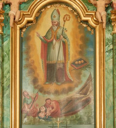 Zdjęcie nr 15: Nastawa ołtarzowa architektoniczna, jednoosiowa, jednokondygnacyjna. Ołtarz prostopadłościenny, drewniany, z drewnianą mensą na niskim cokole, flankowaną po bokach parą kanelowanych pilastrów o kapitelach jońskich. Antependium w formie płyciny marmoryzowanej na kolor różowy w kształcie leżącego prostokąta o owalnych narożach, ujęte w złoconą i wąską ramę. W polu płaskorzeźbione narzędzia Męki Pańskiej (krzyż, włócznia i korona cierniowa). 

Cokół nastawy zryzalitowany, wysoki, zdobiony płycinami z rozetami. Kondygnacja ujęta jest od wewnątrz parą pilastrów o kanelowanych trzonach, a od zewnątrz parą marmoryzowanych na zielono filarów i ustawioną przed nimi parą kolumn o kanelowanych i marmoryzowanych na różowo trzonach. Podpory ustawione są na bazach, mają kompozytowe kapitele i wspierają imposty oraz przerwane nad polem środkowym belkowanie, dekorowane profilowaniem i pasem kostkowania, a także przerwany, wolutowy przyczółek z parą aniołków. Nad środkiem belkowania nad dwóch niewielkich, spiętych spływach wolutowych ustawiona jest niewielkich rozmiarów rzeźbiona grupa przedstawiająca Boga Ojca po prawej i Jezusa Chrystusa po lewej stronie. Nad retabulum, nad wnęką okienną zawieszona jest gołębica Ducha Świętego w dużej promienistej glorii o promieniach, zebranych w wiązki nierównej długości. Retabulum po bokach ujęte jest uszami w formie ażurowej wici z ornamentu rocaille&#039;owego.

W polu środkowym w szerokiej ramie w kształcie stojącego prostokąta, zamkniętego łukiem wklęsło-wypukłym z uskokami umieszczony jest obraz św. Mikołaja oraz św. Barbary na zasuwie. W zwieńczeniu ramy po bokach usadzona jest para aniołków. Poniżej obrazu fantazyjna, architektoniczna oprawa z umieszczonym w polu obrazem św. Teresy z Lisieux. Oprawa w formie niewielkiej, płaskiej aediculi, ustawionej na niskim cokole, flankowanej dwoma pilastrami, zamkniętej łukiem nadwieszonym, na szczycie którego niski cokolik na krucyfiks. Po bokach uszy o fantazyjnym wykroju, z wycięciem w górnej połowie, w dolnej zdobione kratką.

Struktura marmoryzowana jest w odcieniu niebieskim i jasnoróżowym; ornamentyka, profile, ramy i kanelury na pilastrach są złocone.