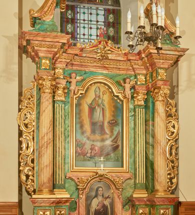 Zdjęcie nr 1: Nastawa ołtarzowa architektoniczna, jednoosiowa, jednokondygnacyjna. Ołtarz prostopadłościenny, drewniany, z drewnianą mensą na niskim cokole, flankowaną po bokach parą kanelowanych pilastrów o kapitelach jońskich. Antependium w formie płyciny marmoryzowanej na kolor różowy w kształcie leżącego prostokąta o owalnych narożach, ujęte w złoconą i wąską ramę. W polu płaskorzeźbione narzędzia Męki Pańskiej (krzyż, włócznia i korona cierniowa). 

Cokół nastawy zryzalitowany, wysoki, zdobiony płycinami z rozetami. Kondygnacja ujęta jest od wewnątrz parą pilastrów o kanelowanych trzonach, a od zewnątrz parą marmoryzowanych na zielono filarów i ustawioną przed nimi parą kolumn o kanelowanych i marmoryzowanych na różowo trzonach. Podpory ustawione są na bazach, mają kompozytowe kapitele i wspierają imposty oraz przerwane nad polem środkowym belkowanie, dekorowane profilowaniem i pasem kostkowania, a także przerwany, wolutowy przyczółek z parą aniołków. Nad środkiem belkowania nad dwóch niewielkich, spiętych spływach wolutowych ustawiona jest niewielkich rozmiarów rzeźbiona grupa przedstawiająca Boga Ojca po prawej i Jezusa Chrystusa po lewej stronie. Nad retabulum, nad wnęką okienną zawieszona jest gołębica Ducha Świętego w dużej promienistej glorii o promieniach, zebranych w wiązki nierównej długości. Retabulum po bokach ujęte jest uszami w formie ażurowej wici z ornamentu rocaille&#039;owego.

W polu środkowym w szerokiej ramie w kształcie stojącego prostokąta, zamkniętego łukiem wklęsło-wypukłym z uskokami umieszczony jest obraz św. Mikołaja oraz św. Barbary na zasuwie. W zwieńczeniu ramy po bokach usadzona jest para aniołków. Poniżej obrazu fantazyjna, architektoniczna oprawa z umieszczonym w polu obrazem św. Teresy z Lisieux. Oprawa w formie niewielkiej, płaskiej aediculi, ustawionej na niskim cokole, flankowanej dwoma pilastrami, zamkniętej łukiem nadwieszonym, na szczycie którego niski cokolik na krucyfiks. Po bokach uszy o fantazyjnym wykroju, z wycięciem w górnej połowie, w dolnej zdobione kratką.

Struktura marmoryzowana jest w odcieniu niebieskim i jasnoróżowym; ornamentyka, profile, ramy i kanelury na pilastrach są złocone.