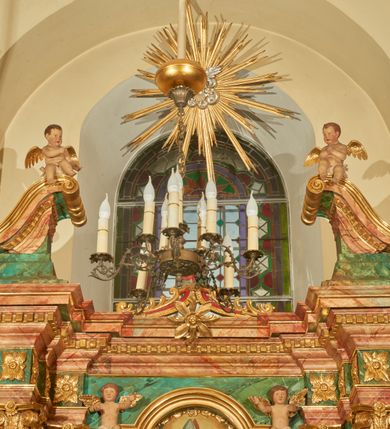 Zdjęcie nr 14: Nastawa ołtarzowa architektoniczna, jednoosiowa, jednokondygnacyjna. Ołtarz prostopadłościenny, drewniany, z drewnianą mensą na niskim cokole, flankowaną po bokach parą kanelowanych pilastrów o kapitelach jońskich. Antependium w formie płyciny marmoryzowanej na kolor różowy w kształcie leżącego prostokąta o owalnych narożach, ujęte w złoconą i wąską ramę. W polu płaskorzeźbione narzędzia Męki Pańskiej (krzyż, włócznia i korona cierniowa). 

Cokół nastawy zryzalitowany, wysoki, zdobiony płycinami z rozetami. Kondygnacja ujęta jest od wewnątrz parą pilastrów o kanelowanych trzonach, a od zewnątrz parą marmoryzowanych na zielono filarów i ustawioną przed nimi parą kolumn o kanelowanych i marmoryzowanych na różowo trzonach. Podpory ustawione są na bazach, mają kompozytowe kapitele i wspierają imposty oraz przerwane nad polem środkowym belkowanie, dekorowane profilowaniem i pasem kostkowania, a także przerwany, wolutowy przyczółek z parą aniołków. Nad środkiem belkowania nad dwóch niewielkich, spiętych spływach wolutowych ustawiona jest niewielkich rozmiarów rzeźbiona grupa przedstawiająca Boga Ojca po prawej i Jezusa Chrystusa po lewej stronie. Nad retabulum, nad wnęką okienną zawieszona jest gołębica Ducha Świętego w dużej promienistej glorii o promieniach, zebranych w wiązki nierównej długości. Retabulum po bokach ujęte jest uszami w formie ażurowej wici z ornamentu rocaille'owego.

W polu środkowym w szerokiej ramie w kształcie stojącego prostokąta, zamkniętego łukiem wklęsło-wypukłym z uskokami umieszczony jest obraz św. Mikołaja oraz św. Barbary na zasuwie. W zwieńczeniu ramy po bokach usadzona jest para aniołków. Poniżej obrazu fantazyjna, architektoniczna oprawa z umieszczonym w polu obrazem św. Teresy z Lisieux. Oprawa w formie niewielkiej, płaskiej aediculi, ustawionej na niskim cokole, flankowanej dwoma pilastrami, zamkniętej łukiem nadwieszonym, na szczycie którego niski cokolik na krucyfiks. Po bokach uszy o fantazyjnym wykroju, z wycięciem w górnej połowie, w dolnej zdobione kratką.

Struktura marmoryzowana jest w odcieniu niebieskim i jasnoróżowym; ornamentyka, profile, ramy i kanelury na pilastrach są złocone.
