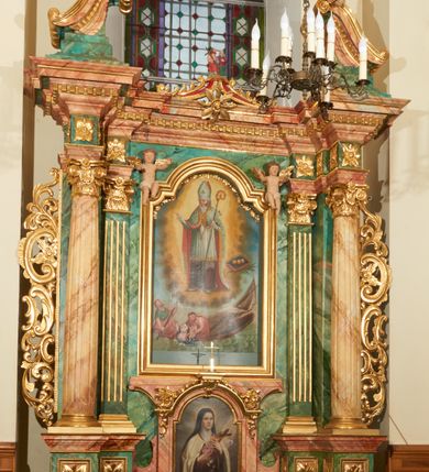 Zdjęcie nr 2: Obraz w kształcie stojącego prostokąta, zamkniętego łukiem wklęsło-wypukłym z uskokami z przedstawieniem św. Mikołaja ratującego rozbitków. Kompozycja dwustrefowa, wielopostaciowa. W górnej strefie, w centrum kompozycji św. Mikołaj ukazany na tle otoku utworzonego z światła i chmur, zwrócony trzy czwarte w prawo, który prawą ręką czyni gest błogosławieństwa, a w lewej trzyma pastorał. Święty ubrany jest w szaty biskupie: sięgającą do ziemi szarą albę, białą rokietę zakończoną złotą lamelką oraz ozdobny płaszcz z czerwoną podszewką i złotym galonem na obrzeżach; na głowie ma infułę. Twarz okolona jest siwą i długą brodą. Po prawej stronie obrazu znajduje się palma męczeństwa i zamknięta księga w ciemnej oprawie, a na niej ułożone rzędem trzy złote kule. W dolnej strefie, w morskiej fali, na zatopionej częściowo łódce z przewróconym, wysokim, białym żaglem widoczna jest grupka tonących ludzi. Po lewej mężczyzna siedzący na końcu łódki, zwrócony trzy czwarte w lewo, z wiosłem w rękach,  z głową skierowaną w stronę św. Mikołaja. Ubrany jest w niebieską suknię z podwiniętym rękawem oraz przerzucony przez lewe ramię czerwony płaszcz, okrywający prawie całą postać.  Twarz ukazana z prawego profilu, włosy i broda siwe, długi i wąski nos. Obok niego mężczyzna ukazany od pasa w górę, ratujący tonącą kobietę i dziecko. Ubrany jest w czerwoną szatę, zarzuconą na prawe ramię i otulającą prawie całą postać. Kobieta do piersi zanurzona w morskiej fali, zwrócona trzy czwarte w prawo, z rękoma wzniesionymi ku górze i głową skierowaną w stronę św. Mikołaja. Ubrana jest w niebieską szatę, odsłaniającą piersi. Twarz okrągła; małe usta; długi i wąski nos, niskie czoło. Włosy ciemne, długie, z przedziałkiem na środku. Na jej lewym ramieniu nagie niemowlę, zwrócone trzy czwarte w prawo, z rękoma wzniesionymi ku górze. Kolorystyka bogata, z przewagą barw błękitu i czerwieni. Rama drewniana, profilowana, złocona, od góry zamknięta łukiem wklęsło-wypukłym z uskokami i ozdobiona girlandą utworzoną z kampanul i kwiatów.