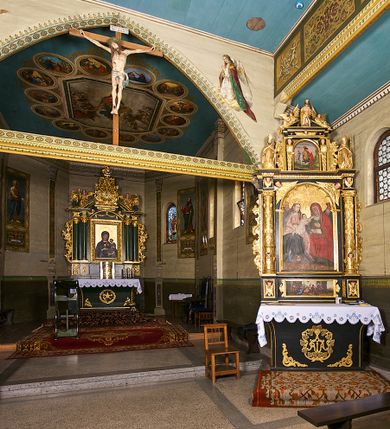 Zdjęcie nr 19: Kościół pomocniczny pw. Wniebowzięcia Najświętszej Marii Panny w Woli Radziszowskiej to niewielkich rozmiarów, orientowana, jednonawowa budowla drewniana, konstrukcji zrębowej, z wieżą w słup. Prezbiterium jest zamknięte wielobocznie, nieco wyższa i szersza nawa ma rzut prostokąta zbliżonego do kwadratu. Do prezbiterium od strony północnej przylega zakrystia, od wschodniej Ogrójec, przy nawie od południa umieszczona została niewielka, prostokątna kruchta. Od zachodu do budowli dostawiona jest czworoboczna wieża o pochyłych ścianach, z podwieszoną izbicą z czworobocznymi występami na przekątnikach; hełm ostrosłupowy z czterema wieżyczkami na występach. W przyziemiu wieży znajduje się kruchta, a dookoła wieży szalowane soboty. Wewnątrz świątyni stropy; w nawie z zaskrzynieniami. Otwór tęczowy ma kształt ostrołuku, belka jest profilowana, na niej umieszczony jest krucyfiks o charakterze późnogotyckim. Chór muzyczny w zachodniej części nawy jest drewniany, wsparty na dwóch kolumnach. Okna nawy i prezbiterium są głównie prostokątne zamknięte półkoliście; z witrażami (z wyjątkiem okna za ołtarzem głównym); ponadto budowlę doświetlają niewielkie okienka prostokątne. Do świątyni prowadzi dwoje drzwi ostrołukowych, trzecie od strony zachodniej przerobiono w 1933 roku. Dachy kościoła są kryte gontem, ponad nawą i kruchtą południową dwuspadowe, nad zakrystią jednospadowy, nad prezbiterium wielopołaciowy. Wieżyczka na sygnaturkę o formach barokowych z glorietą, cebulastym hełmem zwieńczonym kulą oraz krzyżem; kryta blachą. Świątynia usytuowana jest na niewielkiej działce, wydzielonej ogrodzeniem z drewnianych bali, krytym gontem. Bramy wiodące na posesję umieszczono od zachodu oraz w południowo-wschodnim narożniku działki.