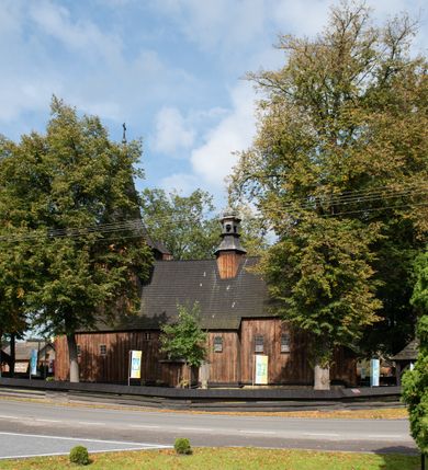 Zdjęcie nr 4: Kościół pomocniczny pw. Wniebowzięcia Najświętszej Marii Panny w Woli Radziszowskiej to niewielkich rozmiarów, orientowana, jednonawowa budowla drewniana, konstrukcji zrębowej, z wieżą w słup. Prezbiterium jest zamknięte wielobocznie, nieco wyższa i szersza nawa ma rzut prostokąta zbliżonego do kwadratu. Do prezbiterium od strony północnej przylega zakrystia, od wschodniej Ogrójec, przy nawie od południa umieszczona została niewielka, prostokątna kruchta. Od zachodu do budowli dostawiona jest czworoboczna wieża o pochyłych ścianach, z podwieszoną izbicą z czworobocznymi występami na przekątnikach; hełm ostrosłupowy z czterema wieżyczkami na występach. W przyziemiu wieży znajduje się kruchta, a dookoła wieży szalowane soboty. Wewnątrz świątyni stropy; w nawie z zaskrzynieniami. Otwór tęczowy ma kształt ostrołuku, belka jest profilowana, na niej umieszczony jest krucyfiks o charakterze późnogotyckim. Chór muzyczny w zachodniej części nawy jest drewniany, wsparty na dwóch kolumnach. Okna nawy i prezbiterium są głównie prostokątne zamknięte półkoliście; z witrażami (z wyjątkiem okna za ołtarzem głównym); ponadto budowlę doświetlają niewielkie okienka prostokątne. Do świątyni prowadzi dwoje drzwi ostrołukowych, trzecie od strony zachodniej przerobiono w 1933 roku. Dachy kościoła są kryte gontem, ponad nawą i kruchtą południową dwuspadowe, nad zakrystią jednospadowy, nad prezbiterium wielopołaciowy. Wieżyczka na sygnaturkę o formach barokowych z glorietą, cebulastym hełmem zwieńczonym kulą oraz krzyżem; kryta blachą. Świątynia usytuowana jest na niewielkiej działce, wydzielonej ogrodzeniem z drewnianych bali, krytym gontem. Bramy wiodące na posesję umieszczono od zachodu oraz w południowo-wschodnim narożniku działki.