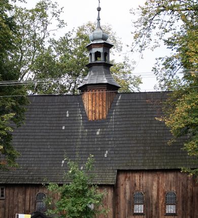 Zdjęcie nr 6: Kościół pomocniczny pw. Wniebowzięcia Najświętszej Marii Panny w Woli Radziszowskiej to niewielkich rozmiarów, orientowana, jednonawowa budowla drewniana, konstrukcji zrębowej, z wieżą w słup. Prezbiterium jest zamknięte wielobocznie, nieco wyższa i szersza nawa ma rzut prostokąta zbliżonego do kwadratu. Do prezbiterium od strony północnej przylega zakrystia, od wschodniej Ogrójec, przy nawie od południa umieszczona została niewielka, prostokątna kruchta. Od zachodu do budowli dostawiona jest czworoboczna wieża o pochyłych ścianach, z podwieszoną izbicą z czworobocznymi występami na przekątnikach; hełm ostrosłupowy z czterema wieżyczkami na występach. W przyziemiu wieży znajduje się kruchta, a dookoła wieży szalowane soboty. Wewnątrz świątyni stropy; w nawie z zaskrzynieniami. Otwór tęczowy ma kształt ostrołuku, belka jest profilowana, na niej umieszczony jest krucyfiks o charakterze późnogotyckim. Chór muzyczny w zachodniej części nawy jest drewniany, wsparty na dwóch kolumnach. Okna nawy i prezbiterium są głównie prostokątne zamknięte półkoliście; z witrażami (z wyjątkiem okna za ołtarzem głównym); ponadto budowlę doświetlają niewielkie okienka prostokątne. Do świątyni prowadzi dwoje drzwi ostrołukowych, trzecie od strony zachodniej przerobiono w 1933 roku. Dachy kościoła są kryte gontem, ponad nawą i kruchtą południową dwuspadowe, nad zakrystią jednospadowy, nad prezbiterium wielopołaciowy. Wieżyczka na sygnaturkę o formach barokowych z glorietą, cebulastym hełmem zwieńczonym kulą oraz krzyżem; kryta blachą. Świątynia usytuowana jest na niewielkiej działce, wydzielonej ogrodzeniem z drewnianych bali, krytym gontem. Bramy wiodące na posesję umieszczono od zachodu oraz w południowo-wschodnim narożniku działki.