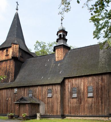 Zdjęcie nr 7: Kościół pomocniczny pw. Wniebowzięcia Najświętszej Marii Panny w Woli Radziszowskiej to niewielkich rozmiarów, orientowana, jednonawowa budowla drewniana, konstrukcji zrębowej, z wieżą w słup. Prezbiterium jest zamknięte wielobocznie, nieco wyższa i szersza nawa ma rzut prostokąta zbliżonego do kwadratu. Do prezbiterium od strony północnej przylega zakrystia, od wschodniej Ogrójec, przy nawie od południa umieszczona została niewielka, prostokątna kruchta. Od zachodu do budowli dostawiona jest czworoboczna wieża o pochyłych ścianach, z podwieszoną izbicą z czworobocznymi występami na przekątnikach; hełm ostrosłupowy z czterema wieżyczkami na występach. W przyziemiu wieży znajduje się kruchta, a dookoła wieży szalowane soboty. Wewnątrz świątyni stropy; w nawie z zaskrzynieniami. Otwór tęczowy ma kształt ostrołuku, belka jest profilowana, na niej umieszczony jest krucyfiks o charakterze późnogotyckim. Chór muzyczny w zachodniej części nawy jest drewniany, wsparty na dwóch kolumnach. Okna nawy i prezbiterium są głównie prostokątne zamknięte półkoliście; z witrażami (z wyjątkiem okna za ołtarzem głównym); ponadto budowlę doświetlają niewielkie okienka prostokątne. Do świątyni prowadzi dwoje drzwi ostrołukowych, trzecie od strony zachodniej przerobiono w 1933 roku. Dachy kościoła są kryte gontem, ponad nawą i kruchtą południową dwuspadowe, nad zakrystią jednospadowy, nad prezbiterium wielopołaciowy. Wieżyczka na sygnaturkę o formach barokowych z glorietą, cebulastym hełmem zwieńczonym kulą oraz krzyżem; kryta blachą. Świątynia usytuowana jest na niewielkiej działce, wydzielonej ogrodzeniem z drewnianych bali, krytym gontem. Bramy wiodące na posesję umieszczono od zachodu oraz w południowo-wschodnim narożniku działki.