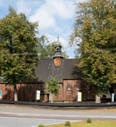 Zdjęcie nr 10: Kościół pomocniczny pw. Wniebowzięcia Najświętszej Marii Panny w Woli Radziszowskiej to niewielkich rozmiarów, orientowana, jednonawowa budowla drewniana, konstrukcji zrębowej, z wieżą w słup. Prezbiterium jest zamknięte wielobocznie, nieco wyższa i szersza nawa ma rzut prostokąta zbliżonego do kwadratu. Do prezbiterium od strony północnej przylega zakrystia, od wschodniej Ogrójec, przy nawie od południa umieszczona została niewielka, prostokątna kruchta. Od zachodu do budowli dostawiona jest czworoboczna wieża o pochyłych ścianach, z podwieszoną izbicą z czworobocznymi występami na przekątnikach; hełm ostrosłupowy z czterema wieżyczkami na występach. W przyziemiu wieży znajduje się kruchta, a dookoła wieży szalowane soboty. Wewnątrz świątyni stropy; w nawie z zaskrzynieniami. Otwór tęczowy ma kształt ostrołuku, belka jest profilowana, na niej umieszczony jest krucyfiks o charakterze późnogotyckim. Chór muzyczny w zachodniej części nawy jest drewniany, wsparty na dwóch kolumnach. Okna nawy i prezbiterium są głównie prostokątne zamknięte półkoliście; z witrażami (z wyjątkiem okna za ołtarzem głównym); ponadto budowlę doświetlają niewielkie okienka prostokątne. Do świątyni prowadzi dwoje drzwi ostrołukowych, trzecie od strony zachodniej przerobiono w 1933 roku. Dachy kościoła są kryte gontem, ponad nawą i kruchtą południową dwuspadowe, nad zakrystią jednospadowy, nad prezbiterium wielopołaciowy. Wieżyczka na sygnaturkę o formach barokowych z glorietą, cebulastym hełmem zwieńczonym kulą oraz krzyżem; kryta blachą. Świątynia usytuowana jest na niewielkiej działce, wydzielonej ogrodzeniem z drewnianych bali, krytym gontem. Bramy wiodące na posesję umieszczono od zachodu oraz w południowo-wschodnim narożniku działki.