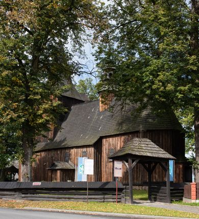 Zdjęcie nr 9: Kościół pomocniczny pw. Wniebowzięcia Najświętszej Marii Panny w Woli Radziszowskiej to niewielkich rozmiarów, orientowana, jednonawowa budowla drewniana, konstrukcji zrębowej, z wieżą w słup. Prezbiterium jest zamknięte wielobocznie, nieco wyższa i szersza nawa ma rzut prostokąta zbliżonego do kwadratu. Do prezbiterium od strony północnej przylega zakrystia, od wschodniej Ogrójec, przy nawie od południa umieszczona została niewielka, prostokątna kruchta. Od zachodu do budowli dostawiona jest czworoboczna wieża o pochyłych ścianach, z podwieszoną izbicą z czworobocznymi występami na przekątnikach; hełm ostrosłupowy z czterema wieżyczkami na występach. W przyziemiu wieży znajduje się kruchta, a dookoła wieży szalowane soboty. Wewnątrz świątyni stropy; w nawie z zaskrzynieniami. Otwór tęczowy ma kształt ostrołuku, belka jest profilowana, na niej umieszczony jest krucyfiks o charakterze późnogotyckim. Chór muzyczny w zachodniej części nawy jest drewniany, wsparty na dwóch kolumnach. Okna nawy i prezbiterium są głównie prostokątne zamknięte półkoliście; z witrażami (z wyjątkiem okna za ołtarzem głównym); ponadto budowlę doświetlają niewielkie okienka prostokątne. Do świątyni prowadzi dwoje drzwi ostrołukowych, trzecie od strony zachodniej przerobiono w 1933 roku. Dachy kościoła są kryte gontem, ponad nawą i kruchtą południową dwuspadowe, nad zakrystią jednospadowy, nad prezbiterium wielopołaciowy. Wieżyczka na sygnaturkę o formach barokowych z glorietą, cebulastym hełmem zwieńczonym kulą oraz krzyżem; kryta blachą. Świątynia usytuowana jest na niewielkiej działce, wydzielonej ogrodzeniem z drewnianych bali, krytym gontem. Bramy wiodące na posesję umieszczono od zachodu oraz w południowo-wschodnim narożniku działki.