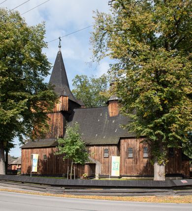 Zdjęcie nr 12: Kościół pomocniczny pw. Wniebowzięcia Najświętszej Marii Panny w Woli Radziszowskiej to niewielkich rozmiarów, orientowana, jednonawowa budowla drewniana, konstrukcji zrębowej, z wieżą w słup. Prezbiterium jest zamknięte wielobocznie, nieco wyższa i szersza nawa ma rzut prostokąta zbliżonego do kwadratu. Do prezbiterium od strony północnej przylega zakrystia, od wschodniej Ogrójec, przy nawie od południa umieszczona została niewielka, prostokątna kruchta. Od zachodu do budowli dostawiona jest czworoboczna wieża o pochyłych ścianach, z podwieszoną izbicą z czworobocznymi występami na przekątnikach; hełm ostrosłupowy z czterema wieżyczkami na występach. W przyziemiu wieży znajduje się kruchta, a dookoła wieży szalowane soboty. Wewnątrz świątyni stropy; w nawie z zaskrzynieniami. Otwór tęczowy ma kształt ostrołuku, belka jest profilowana, na niej umieszczony jest krucyfiks o charakterze późnogotyckim. Chór muzyczny w zachodniej części nawy jest drewniany, wsparty na dwóch kolumnach. Okna nawy i prezbiterium są głównie prostokątne zamknięte półkoliście; z witrażami (z wyjątkiem okna za ołtarzem głównym); ponadto budowlę doświetlają niewielkie okienka prostokątne. Do świątyni prowadzi dwoje drzwi ostrołukowych, trzecie od strony zachodniej przerobiono w 1933 roku. Dachy kościoła są kryte gontem, ponad nawą i kruchtą południową dwuspadowe, nad zakrystią jednospadowy, nad prezbiterium wielopołaciowy. Wieżyczka na sygnaturkę o formach barokowych z glorietą, cebulastym hełmem zwieńczonym kulą oraz krzyżem; kryta blachą. Świątynia usytuowana jest na niewielkiej działce, wydzielonej ogrodzeniem z drewnianych bali, krytym gontem. Bramy wiodące na posesję umieszczono od zachodu oraz w południowo-wschodnim narożniku działki.