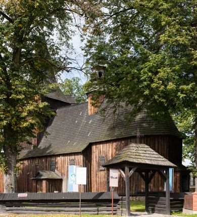 Zdjęcie nr 11: Kościół pomocniczny pw. Wniebowzięcia Najświętszej Marii Panny w Woli Radziszowskiej to niewielkich rozmiarów, orientowana, jednonawowa budowla drewniana, konstrukcji zrębowej, z wieżą w słup. Prezbiterium jest zamknięte wielobocznie, nieco wyższa i szersza nawa ma rzut prostokąta zbliżonego do kwadratu. Do prezbiterium od strony północnej przylega zakrystia, od wschodniej Ogrójec, przy nawie od południa umieszczona została niewielka, prostokątna kruchta. Od zachodu do budowli dostawiona jest czworoboczna wieża o pochyłych ścianach, z podwieszoną izbicą z czworobocznymi występami na przekątnikach; hełm ostrosłupowy z czterema wieżyczkami na występach. W przyziemiu wieży znajduje się kruchta, a dookoła wieży szalowane soboty. Wewnątrz świątyni stropy; w nawie z zaskrzynieniami. Otwór tęczowy ma kształt ostrołuku, belka jest profilowana, na niej umieszczony jest krucyfiks o charakterze późnogotyckim. Chór muzyczny w zachodniej części nawy jest drewniany, wsparty na dwóch kolumnach. Okna nawy i prezbiterium są głównie prostokątne zamknięte półkoliście; z witrażami (z wyjątkiem okna za ołtarzem głównym); ponadto budowlę doświetlają niewielkie okienka prostokątne. Do świątyni prowadzi dwoje drzwi ostrołukowych, trzecie od strony zachodniej przerobiono w 1933 roku. Dachy kościoła są kryte gontem, ponad nawą i kruchtą południową dwuspadowe, nad zakrystią jednospadowy, nad prezbiterium wielopołaciowy. Wieżyczka na sygnaturkę o formach barokowych z glorietą, cebulastym hełmem zwieńczonym kulą oraz krzyżem; kryta blachą. Świątynia usytuowana jest na niewielkiej działce, wydzielonej ogrodzeniem z drewnianych bali, krytym gontem. Bramy wiodące na posesję umieszczono od zachodu oraz w południowo-wschodnim narożniku działki.