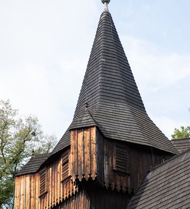 Zdjęcie nr 13: Kościół pomocniczny pw. Wniebowzięcia Najświętszej Marii Panny w Woli Radziszowskiej to niewielkich rozmiarów, orientowana, jednonawowa budowla drewniana, konstrukcji zrębowej, z wieżą w słup. Prezbiterium jest zamknięte wielobocznie, nieco wyższa i szersza nawa ma rzut prostokąta zbliżonego do kwadratu. Do prezbiterium od strony północnej przylega zakrystia, od wschodniej Ogrójec, przy nawie od południa umieszczona została niewielka, prostokątna kruchta. Od zachodu do budowli dostawiona jest czworoboczna wieża o pochyłych ścianach, z podwieszoną izbicą z czworobocznymi występami na przekątnikach; hełm ostrosłupowy z czterema wieżyczkami na występach. W przyziemiu wieży znajduje się kruchta, a dookoła wieży szalowane soboty. Wewnątrz świątyni stropy; w nawie z zaskrzynieniami. Otwór tęczowy ma kształt ostrołuku, belka jest profilowana, na niej umieszczony jest krucyfiks o charakterze późnogotyckim. Chór muzyczny w zachodniej części nawy jest drewniany, wsparty na dwóch kolumnach. Okna nawy i prezbiterium są głównie prostokątne zamknięte półkoliście; z witrażami (z wyjątkiem okna za ołtarzem głównym); ponadto budowlę doświetlają niewielkie okienka prostokątne. Do świątyni prowadzi dwoje drzwi ostrołukowych, trzecie od strony zachodniej przerobiono w 1933 roku. Dachy kościoła są kryte gontem, ponad nawą i kruchtą południową dwuspadowe, nad zakrystią jednospadowy, nad prezbiterium wielopołaciowy. Wieżyczka na sygnaturkę o formach barokowych z glorietą, cebulastym hełmem zwieńczonym kulą oraz krzyżem; kryta blachą. Świątynia usytuowana jest na niewielkiej działce, wydzielonej ogrodzeniem z drewnianych bali, krytym gontem. Bramy wiodące na posesję umieszczono od zachodu oraz w południowo-wschodnim narożniku działki.