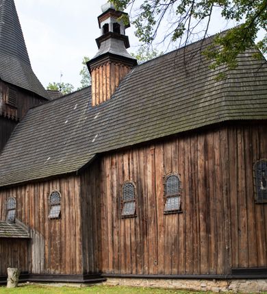 Zdjęcie nr 14: Kościół pomocniczny pw. Wniebowzięcia Najświętszej Marii Panny w Woli Radziszowskiej to niewielkich rozmiarów, orientowana, jednonawowa budowla drewniana, konstrukcji zrębowej, z wieżą w słup. Prezbiterium jest zamknięte wielobocznie, nieco wyższa i szersza nawa ma rzut prostokąta zbliżonego do kwadratu. Do prezbiterium od strony północnej przylega zakrystia, od wschodniej Ogrójec, przy nawie od południa umieszczona została niewielka, prostokątna kruchta. Od zachodu do budowli dostawiona jest czworoboczna wieża o pochyłych ścianach, z podwieszoną izbicą z czworobocznymi występami na przekątnikach; hełm ostrosłupowy z czterema wieżyczkami na występach. W przyziemiu wieży znajduje się kruchta, a dookoła wieży szalowane soboty. Wewnątrz świątyni stropy; w nawie z zaskrzynieniami. Otwór tęczowy ma kształt ostrołuku, belka jest profilowana, na niej umieszczony jest krucyfiks o charakterze późnogotyckim. Chór muzyczny w zachodniej części nawy jest drewniany, wsparty na dwóch kolumnach. Okna nawy i prezbiterium są głównie prostokątne zamknięte półkoliście; z witrażami (z wyjątkiem okna za ołtarzem głównym); ponadto budowlę doświetlają niewielkie okienka prostokątne. Do świątyni prowadzi dwoje drzwi ostrołukowych, trzecie od strony zachodniej przerobiono w 1933 roku. Dachy kościoła są kryte gontem, ponad nawą i kruchtą południową dwuspadowe, nad zakrystią jednospadowy, nad prezbiterium wielopołaciowy. Wieżyczka na sygnaturkę o formach barokowych z glorietą, cebulastym hełmem zwieńczonym kulą oraz krzyżem; kryta blachą. Świątynia usytuowana jest na niewielkiej działce, wydzielonej ogrodzeniem z drewnianych bali, krytym gontem. Bramy wiodące na posesję umieszczono od zachodu oraz w południowo-wschodnim narożniku działki.