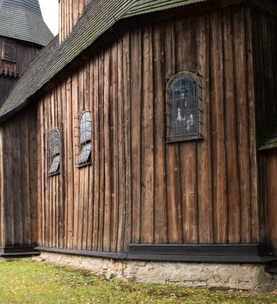 Zdjęcie nr 15: Kościół pomocniczny pw. Wniebowzięcia Najświętszej Marii Panny w Woli Radziszowskiej to niewielkich rozmiarów, orientowana, jednonawowa budowla drewniana, konstrukcji zrębowej, z wieżą w słup. Prezbiterium jest zamknięte wielobocznie, nieco wyższa i szersza nawa ma rzut prostokąta zbliżonego do kwadratu. Do prezbiterium od strony północnej przylega zakrystia, od wschodniej Ogrójec, przy nawie od południa umieszczona została niewielka, prostokątna kruchta. Od zachodu do budowli dostawiona jest czworoboczna wieża o pochyłych ścianach, z podwieszoną izbicą z czworobocznymi występami na przekątnikach; hełm ostrosłupowy z czterema wieżyczkami na występach. W przyziemiu wieży znajduje się kruchta, a dookoła wieży szalowane soboty. Wewnątrz świątyni stropy; w nawie z zaskrzynieniami. Otwór tęczowy ma kształt ostrołuku, belka jest profilowana, na niej umieszczony jest krucyfiks o charakterze późnogotyckim. Chór muzyczny w zachodniej części nawy jest drewniany, wsparty na dwóch kolumnach. Okna nawy i prezbiterium są głównie prostokątne zamknięte półkoliście; z witrażami (z wyjątkiem okna za ołtarzem głównym); ponadto budowlę doświetlają niewielkie okienka prostokątne. Do świątyni prowadzi dwoje drzwi ostrołukowych, trzecie od strony zachodniej przerobiono w 1933 roku. Dachy kościoła są kryte gontem, ponad nawą i kruchtą południową dwuspadowe, nad zakrystią jednospadowy, nad prezbiterium wielopołaciowy. Wieżyczka na sygnaturkę o formach barokowych z glorietą, cebulastym hełmem zwieńczonym kulą oraz krzyżem; kryta blachą. Świątynia usytuowana jest na niewielkiej działce, wydzielonej ogrodzeniem z drewnianych bali, krytym gontem. Bramy wiodące na posesję umieszczono od zachodu oraz w południowo-wschodnim narożniku działki.