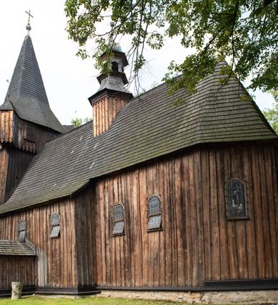 Zdjęcie nr 16: Kościół pomocniczny pw. Wniebowzięcia Najświętszej Marii Panny w Woli Radziszowskiej to niewielkich rozmiarów, orientowana, jednonawowa budowla drewniana, konstrukcji zrębowej, z wieżą w słup. Prezbiterium jest zamknięte wielobocznie, nieco wyższa i szersza nawa ma rzut prostokąta zbliżonego do kwadratu. Do prezbiterium od strony północnej przylega zakrystia, od wschodniej Ogrójec, przy nawie od południa umieszczona została niewielka, prostokątna kruchta. Od zachodu do budowli dostawiona jest czworoboczna wieża o pochyłych ścianach, z podwieszoną izbicą z czworobocznymi występami na przekątnikach; hełm ostrosłupowy z czterema wieżyczkami na występach. W przyziemiu wieży znajduje się kruchta, a dookoła wieży szalowane soboty. Wewnątrz świątyni stropy; w nawie z zaskrzynieniami. Otwór tęczowy ma kształt ostrołuku, belka jest profilowana, na niej umieszczony jest krucyfiks o charakterze późnogotyckim. Chór muzyczny w zachodniej części nawy jest drewniany, wsparty na dwóch kolumnach. Okna nawy i prezbiterium są głównie prostokątne zamknięte półkoliście; z witrażami (z wyjątkiem okna za ołtarzem głównym); ponadto budowlę doświetlają niewielkie okienka prostokątne. Do świątyni prowadzi dwoje drzwi ostrołukowych, trzecie od strony zachodniej przerobiono w 1933 roku. Dachy kościoła są kryte gontem, ponad nawą i kruchtą południową dwuspadowe, nad zakrystią jednospadowy, nad prezbiterium wielopołaciowy. Wieżyczka na sygnaturkę o formach barokowych z glorietą, cebulastym hełmem zwieńczonym kulą oraz krzyżem; kryta blachą. Świątynia usytuowana jest na niewielkiej działce, wydzielonej ogrodzeniem z drewnianych bali, krytym gontem. Bramy wiodące na posesję umieszczono od zachodu oraz w południowo-wschodnim narożniku działki.