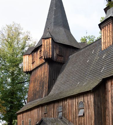 Zdjęcie nr 17: Kościół pomocniczny pw. Wniebowzięcia Najświętszej Marii Panny w Woli Radziszowskiej to niewielkich rozmiarów, orientowana, jednonawowa budowla drewniana, konstrukcji zrębowej, z wieżą w słup. Prezbiterium jest zamknięte wielobocznie, nieco wyższa i szersza nawa ma rzut prostokąta zbliżonego do kwadratu. Do prezbiterium od strony północnej przylega zakrystia, od wschodniej Ogrójec, przy nawie od południa umieszczona została niewielka, prostokątna kruchta. Od zachodu do budowli dostawiona jest czworoboczna wieża o pochyłych ścianach, z podwieszoną izbicą z czworobocznymi występami na przekątnikach; hełm ostrosłupowy z czterema wieżyczkami na występach. W przyziemiu wieży znajduje się kruchta, a dookoła wieży szalowane soboty. Wewnątrz świątyni stropy; w nawie z zaskrzynieniami. Otwór tęczowy ma kształt ostrołuku, belka jest profilowana, na niej umieszczony jest krucyfiks o charakterze późnogotyckim. Chór muzyczny w zachodniej części nawy jest drewniany, wsparty na dwóch kolumnach. Okna nawy i prezbiterium są głównie prostokątne zamknięte półkoliście; z witrażami (z wyjątkiem okna za ołtarzem głównym); ponadto budowlę doświetlają niewielkie okienka prostokątne. Do świątyni prowadzi dwoje drzwi ostrołukowych, trzecie od strony zachodniej przerobiono w 1933 roku. Dachy kościoła są kryte gontem, ponad nawą i kruchtą południową dwuspadowe, nad zakrystią jednospadowy, nad prezbiterium wielopołaciowy. Wieżyczka na sygnaturkę o formach barokowych z glorietą, cebulastym hełmem zwieńczonym kulą oraz krzyżem; kryta blachą. Świątynia usytuowana jest na niewielkiej działce, wydzielonej ogrodzeniem z drewnianych bali, krytym gontem. Bramy wiodące na posesję umieszczono od zachodu oraz w południowo-wschodnim narożniku działki.