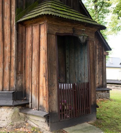 Zdjęcie nr 18: Kościół pomocniczny pw. Wniebowzięcia Najświętszej Marii Panny w Woli Radziszowskiej to niewielkich rozmiarów, orientowana, jednonawowa budowla drewniana, konstrukcji zrębowej, z wieżą w słup. Prezbiterium jest zamknięte wielobocznie, nieco wyższa i szersza nawa ma rzut prostokąta zbliżonego do kwadratu. Do prezbiterium od strony północnej przylega zakrystia, od wschodniej Ogrójec, przy nawie od południa umieszczona została niewielka, prostokątna kruchta. Od zachodu do budowli dostawiona jest czworoboczna wieża o pochyłych ścianach, z podwieszoną izbicą z czworobocznymi występami na przekątnikach; hełm ostrosłupowy z czterema wieżyczkami na występach. W przyziemiu wieży znajduje się kruchta, a dookoła wieży szalowane soboty. Wewnątrz świątyni stropy; w nawie z zaskrzynieniami. Otwór tęczowy ma kształt ostrołuku, belka jest profilowana, na niej umieszczony jest krucyfiks o charakterze późnogotyckim. Chór muzyczny w zachodniej części nawy jest drewniany, wsparty na dwóch kolumnach. Okna nawy i prezbiterium są głównie prostokątne zamknięte półkoliście; z witrażami (z wyjątkiem okna za ołtarzem głównym); ponadto budowlę doświetlają niewielkie okienka prostokątne. Do świątyni prowadzi dwoje drzwi ostrołukowych, trzecie od strony zachodniej przerobiono w 1933 roku. Dachy kościoła są kryte gontem, ponad nawą i kruchtą południową dwuspadowe, nad zakrystią jednospadowy, nad prezbiterium wielopołaciowy. Wieżyczka na sygnaturkę o formach barokowych z glorietą, cebulastym hełmem zwieńczonym kulą oraz krzyżem; kryta blachą. Świątynia usytuowana jest na niewielkiej działce, wydzielonej ogrodzeniem z drewnianych bali, krytym gontem. Bramy wiodące na posesję umieszczono od zachodu oraz w południowo-wschodnim narożniku działki.