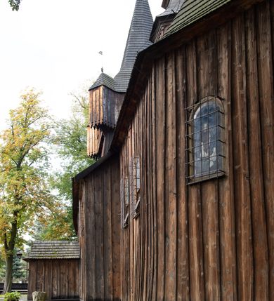 Zdjęcie nr 21: Kościół pomocniczny pw. Wniebowzięcia Najświętszej Marii Panny w Woli Radziszowskiej to niewielkich rozmiarów, orientowana, jednonawowa budowla drewniana, konstrukcji zrębowej, z wieżą w słup. Prezbiterium jest zamknięte wielobocznie, nieco wyższa i szersza nawa ma rzut prostokąta zbliżonego do kwadratu. Do prezbiterium od strony północnej przylega zakrystia, od wschodniej Ogrójec, przy nawie od południa umieszczona została niewielka, prostokątna kruchta. Od zachodu do budowli dostawiona jest czworoboczna wieża o pochyłych ścianach, z podwieszoną izbicą z czworobocznymi występami na przekątnikach; hełm ostrosłupowy z czterema wieżyczkami na występach. W przyziemiu wieży znajduje się kruchta, a dookoła wieży szalowane soboty. Wewnątrz świątyni stropy; w nawie z zaskrzynieniami. Otwór tęczowy ma kształt ostrołuku, belka jest profilowana, na niej umieszczony jest krucyfiks o charakterze późnogotyckim. Chór muzyczny w zachodniej części nawy jest drewniany, wsparty na dwóch kolumnach. Okna nawy i prezbiterium są głównie prostokątne zamknięte półkoliście; z witrażami (z wyjątkiem okna za ołtarzem głównym); ponadto budowlę doświetlają niewielkie okienka prostokątne. Do świątyni prowadzi dwoje drzwi ostrołukowych, trzecie od strony zachodniej przerobiono w 1933 roku. Dachy kościoła są kryte gontem, ponad nawą i kruchtą południową dwuspadowe, nad zakrystią jednospadowy, nad prezbiterium wielopołaciowy. Wieżyczka na sygnaturkę o formach barokowych z glorietą, cebulastym hełmem zwieńczonym kulą oraz krzyżem; kryta blachą. Świątynia usytuowana jest na niewielkiej działce, wydzielonej ogrodzeniem z drewnianych bali, krytym gontem. Bramy wiodące na posesję umieszczono od zachodu oraz w południowo-wschodnim narożniku działki.