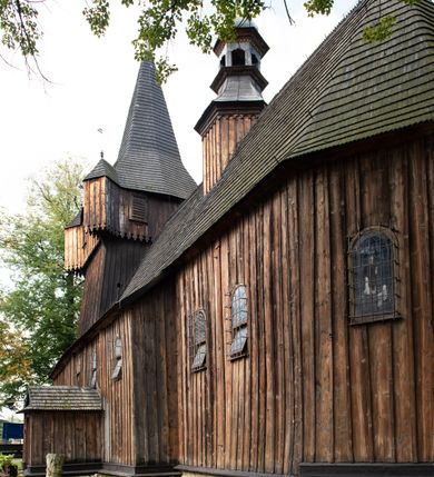 Zdjęcie nr 20: Kościół pomocniczny pw. Wniebowzięcia Najświętszej Marii Panny w Woli Radziszowskiej to niewielkich rozmiarów, orientowana, jednonawowa budowla drewniana, konstrukcji zrębowej, z wieżą w słup. Prezbiterium jest zamknięte wielobocznie, nieco wyższa i szersza nawa ma rzut prostokąta zbliżonego do kwadratu. Do prezbiterium od strony północnej przylega zakrystia, od wschodniej Ogrójec, przy nawie od południa umieszczona została niewielka, prostokątna kruchta. Od zachodu do budowli dostawiona jest czworoboczna wieża o pochyłych ścianach, z podwieszoną izbicą z czworobocznymi występami na przekątnikach; hełm ostrosłupowy z czterema wieżyczkami na występach. W przyziemiu wieży znajduje się kruchta, a dookoła wieży szalowane soboty. Wewnątrz świątyni stropy; w nawie z zaskrzynieniami. Otwór tęczowy ma kształt ostrołuku, belka jest profilowana, na niej umieszczony jest krucyfiks o charakterze późnogotyckim. Chór muzyczny w zachodniej części nawy jest drewniany, wsparty na dwóch kolumnach. Okna nawy i prezbiterium są głównie prostokątne zamknięte półkoliście; z witrażami (z wyjątkiem okna za ołtarzem głównym); ponadto budowlę doświetlają niewielkie okienka prostokątne. Do świątyni prowadzi dwoje drzwi ostrołukowych, trzecie od strony zachodniej przerobiono w 1933 roku. Dachy kościoła są kryte gontem, ponad nawą i kruchtą południową dwuspadowe, nad zakrystią jednospadowy, nad prezbiterium wielopołaciowy. Wieżyczka na sygnaturkę o formach barokowych z glorietą, cebulastym hełmem zwieńczonym kulą oraz krzyżem; kryta blachą. Świątynia usytuowana jest na niewielkiej działce, wydzielonej ogrodzeniem z drewnianych bali, krytym gontem. Bramy wiodące na posesję umieszczono od zachodu oraz w południowo-wschodnim narożniku działki.