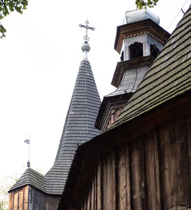 Zdjęcie nr 23: Kościół pomocniczny pw. Wniebowzięcia Najświętszej Marii Panny w Woli Radziszowskiej to niewielkich rozmiarów, orientowana, jednonawowa budowla drewniana, konstrukcji zrębowej, z wieżą w słup. Prezbiterium jest zamknięte wielobocznie, nieco wyższa i szersza nawa ma rzut prostokąta zbliżonego do kwadratu. Do prezbiterium od strony północnej przylega zakrystia, od wschodniej Ogrójec, przy nawie od południa umieszczona została niewielka, prostokątna kruchta. Od zachodu do budowli dostawiona jest czworoboczna wieża o pochyłych ścianach, z podwieszoną izbicą z czworobocznymi występami na przekątnikach; hełm ostrosłupowy z czterema wieżyczkami na występach. W przyziemiu wieży znajduje się kruchta, a dookoła wieży szalowane soboty. Wewnątrz świątyni stropy; w nawie z zaskrzynieniami. Otwór tęczowy ma kształt ostrołuku, belka jest profilowana, na niej umieszczony jest krucyfiks o charakterze późnogotyckim. Chór muzyczny w zachodniej części nawy jest drewniany, wsparty na dwóch kolumnach. Okna nawy i prezbiterium są głównie prostokątne zamknięte półkoliście; z witrażami (z wyjątkiem okna za ołtarzem głównym); ponadto budowlę doświetlają niewielkie okienka prostokątne. Do świątyni prowadzi dwoje drzwi ostrołukowych, trzecie od strony zachodniej przerobiono w 1933 roku. Dachy kościoła są kryte gontem, ponad nawą i kruchtą południową dwuspadowe, nad zakrystią jednospadowy, nad prezbiterium wielopołaciowy. Wieżyczka na sygnaturkę o formach barokowych z glorietą, cebulastym hełmem zwieńczonym kulą oraz krzyżem; kryta blachą. Świątynia usytuowana jest na niewielkiej działce, wydzielonej ogrodzeniem z drewnianych bali, krytym gontem. Bramy wiodące na posesję umieszczono od zachodu oraz w południowo-wschodnim narożniku działki.