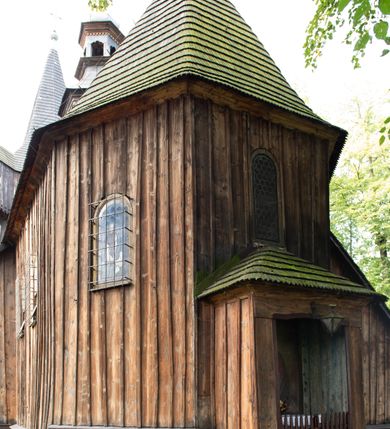 Zdjęcie nr 24: Kościół pomocniczny pw. Wniebowzięcia Najświętszej Marii Panny w Woli Radziszowskiej to niewielkich rozmiarów, orientowana, jednonawowa budowla drewniana, konstrukcji zrębowej, z wieżą w słup. Prezbiterium jest zamknięte wielobocznie, nieco wyższa i szersza nawa ma rzut prostokąta zbliżonego do kwadratu. Do prezbiterium od strony północnej przylega zakrystia, od wschodniej Ogrójec, przy nawie od południa umieszczona została niewielka, prostokątna kruchta. Od zachodu do budowli dostawiona jest czworoboczna wieża o pochyłych ścianach, z podwieszoną izbicą z czworobocznymi występami na przekątnikach; hełm ostrosłupowy z czterema wieżyczkami na występach. W przyziemiu wieży znajduje się kruchta, a dookoła wieży szalowane soboty. Wewnątrz świątyni stropy; w nawie z zaskrzynieniami. Otwór tęczowy ma kształt ostrołuku, belka jest profilowana, na niej umieszczony jest krucyfiks o charakterze późnogotyckim. Chór muzyczny w zachodniej części nawy jest drewniany, wsparty na dwóch kolumnach. Okna nawy i prezbiterium są głównie prostokątne zamknięte półkoliście; z witrażami (z wyjątkiem okna za ołtarzem głównym); ponadto budowlę doświetlają niewielkie okienka prostokątne. Do świątyni prowadzi dwoje drzwi ostrołukowych, trzecie od strony zachodniej przerobiono w 1933 roku. Dachy kościoła są kryte gontem, ponad nawą i kruchtą południową dwuspadowe, nad zakrystią jednospadowy, nad prezbiterium wielopołaciowy. Wieżyczka na sygnaturkę o formach barokowych z glorietą, cebulastym hełmem zwieńczonym kulą oraz krzyżem; kryta blachą. Świątynia usytuowana jest na niewielkiej działce, wydzielonej ogrodzeniem z drewnianych bali, krytym gontem. Bramy wiodące na posesję umieszczono od zachodu oraz w południowo-wschodnim narożniku działki.