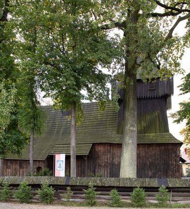 Zdjęcie nr 27: Kościół pomocniczny pw. Wniebowzięcia Najświętszej Marii Panny w Woli Radziszowskiej to niewielkich rozmiarów, orientowana, jednonawowa budowla drewniana, konstrukcji zrębowej, z wieżą w słup. Prezbiterium jest zamknięte wielobocznie, nieco wyższa i szersza nawa ma rzut prostokąta zbliżonego do kwadratu. Do prezbiterium od strony północnej przylega zakrystia, od wschodniej Ogrójec, przy nawie od południa umieszczona została niewielka, prostokątna kruchta. Od zachodu do budowli dostawiona jest czworoboczna wieża o pochyłych ścianach, z podwieszoną izbicą z czworobocznymi występami na przekątnikach; hełm ostrosłupowy z czterema wieżyczkami na występach. W przyziemiu wieży znajduje się kruchta, a dookoła wieży szalowane soboty. Wewnątrz świątyni stropy; w nawie z zaskrzynieniami. Otwór tęczowy ma kształt ostrołuku, belka jest profilowana, na niej umieszczony jest krucyfiks o charakterze późnogotyckim. Chór muzyczny w zachodniej części nawy jest drewniany, wsparty na dwóch kolumnach. Okna nawy i prezbiterium są głównie prostokątne zamknięte półkoliście; z witrażami (z wyjątkiem okna za ołtarzem głównym); ponadto budowlę doświetlają niewielkie okienka prostokątne. Do świątyni prowadzi dwoje drzwi ostrołukowych, trzecie od strony zachodniej przerobiono w 1933 roku. Dachy kościoła są kryte gontem, ponad nawą i kruchtą południową dwuspadowe, nad zakrystią jednospadowy, nad prezbiterium wielopołaciowy. Wieżyczka na sygnaturkę o formach barokowych z glorietą, cebulastym hełmem zwieńczonym kulą oraz krzyżem; kryta blachą. Świątynia usytuowana jest na niewielkiej działce, wydzielonej ogrodzeniem z drewnianych bali, krytym gontem. Bramy wiodące na posesję umieszczono od zachodu oraz w południowo-wschodnim narożniku działki.