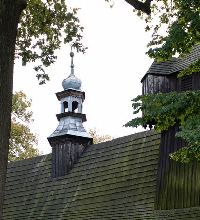 Zdjęcie nr 29: Kościół pomocniczny pw. Wniebowzięcia Najświętszej Marii Panny w Woli Radziszowskiej to niewielkich rozmiarów, orientowana, jednonawowa budowla drewniana, konstrukcji zrębowej, z wieżą w słup. Prezbiterium jest zamknięte wielobocznie, nieco wyższa i szersza nawa ma rzut prostokąta zbliżonego do kwadratu. Do prezbiterium od strony północnej przylega zakrystia, od wschodniej Ogrójec, przy nawie od południa umieszczona została niewielka, prostokątna kruchta. Od zachodu do budowli dostawiona jest czworoboczna wieża o pochyłych ścianach, z podwieszoną izbicą z czworobocznymi występami na przekątnikach; hełm ostrosłupowy z czterema wieżyczkami na występach. W przyziemiu wieży znajduje się kruchta, a dookoła wieży szalowane soboty. Wewnątrz świątyni stropy; w nawie z zaskrzynieniami. Otwór tęczowy ma kształt ostrołuku, belka jest profilowana, na niej umieszczony jest krucyfiks o charakterze późnogotyckim. Chór muzyczny w zachodniej części nawy jest drewniany, wsparty na dwóch kolumnach. Okna nawy i prezbiterium są głównie prostokątne zamknięte półkoliście; z witrażami (z wyjątkiem okna za ołtarzem głównym); ponadto budowlę doświetlają niewielkie okienka prostokątne. Do świątyni prowadzi dwoje drzwi ostrołukowych, trzecie od strony zachodniej przerobiono w 1933 roku. Dachy kościoła są kryte gontem, ponad nawą i kruchtą południową dwuspadowe, nad zakrystią jednospadowy, nad prezbiterium wielopołaciowy. Wieżyczka na sygnaturkę o formach barokowych z glorietą, cebulastym hełmem zwieńczonym kulą oraz krzyżem; kryta blachą. Świątynia usytuowana jest na niewielkiej działce, wydzielonej ogrodzeniem z drewnianych bali, krytym gontem. Bramy wiodące na posesję umieszczono od zachodu oraz w południowo-wschodnim narożniku działki.