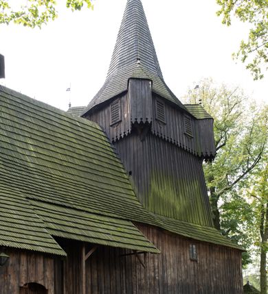 Zdjęcie nr 31: Kościół pomocniczny pw. Wniebowzięcia Najświętszej Marii Panny w Woli Radziszowskiej to niewielkich rozmiarów, orientowana, jednonawowa budowla drewniana, konstrukcji zrębowej, z wieżą w słup. Prezbiterium jest zamknięte wielobocznie, nieco wyższa i szersza nawa ma rzut prostokąta zbliżonego do kwadratu. Do prezbiterium od strony północnej przylega zakrystia, od wschodniej Ogrójec, przy nawie od południa umieszczona została niewielka, prostokątna kruchta. Od zachodu do budowli dostawiona jest czworoboczna wieża o pochyłych ścianach, z podwieszoną izbicą z czworobocznymi występami na przekątnikach; hełm ostrosłupowy z czterema wieżyczkami na występach. W przyziemiu wieży znajduje się kruchta, a dookoła wieży szalowane soboty. Wewnątrz świątyni stropy; w nawie z zaskrzynieniami. Otwór tęczowy ma kształt ostrołuku, belka jest profilowana, na niej umieszczony jest krucyfiks o charakterze późnogotyckim. Chór muzyczny w zachodniej części nawy jest drewniany, wsparty na dwóch kolumnach. Okna nawy i prezbiterium są głównie prostokątne zamknięte półkoliście; z witrażami (z wyjątkiem okna za ołtarzem głównym); ponadto budowlę doświetlają niewielkie okienka prostokątne. Do świątyni prowadzi dwoje drzwi ostrołukowych, trzecie od strony zachodniej przerobiono w 1933 roku. Dachy kościoła są kryte gontem, ponad nawą i kruchtą południową dwuspadowe, nad zakrystią jednospadowy, nad prezbiterium wielopołaciowy. Wieżyczka na sygnaturkę o formach barokowych z glorietą, cebulastym hełmem zwieńczonym kulą oraz krzyżem; kryta blachą. Świątynia usytuowana jest na niewielkiej działce, wydzielonej ogrodzeniem z drewnianych bali, krytym gontem. Bramy wiodące na posesję umieszczono od zachodu oraz w południowo-wschodnim narożniku działki.