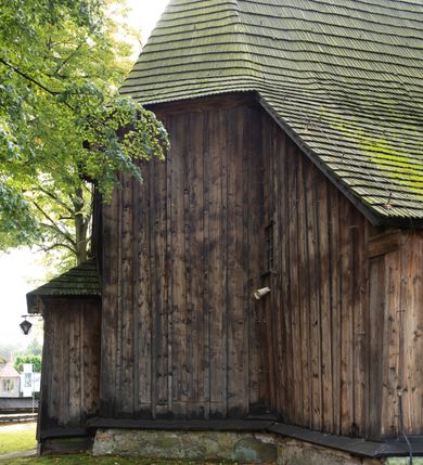 Zdjęcie nr 28: Kościół pomocniczny pw. Wniebowzięcia Najświętszej Marii Panny w Woli Radziszowskiej to niewielkich rozmiarów, orientowana, jednonawowa budowla drewniana, konstrukcji zrębowej, z wieżą w słup. Prezbiterium jest zamknięte wielobocznie, nieco wyższa i szersza nawa ma rzut prostokąta zbliżonego do kwadratu. Do prezbiterium od strony północnej przylega zakrystia, od wschodniej Ogrójec, przy nawie od południa umieszczona została niewielka, prostokątna kruchta. Od zachodu do budowli dostawiona jest czworoboczna wieża o pochyłych ścianach, z podwieszoną izbicą z czworobocznymi występami na przekątnikach; hełm ostrosłupowy z czterema wieżyczkami na występach. W przyziemiu wieży znajduje się kruchta, a dookoła wieży szalowane soboty. Wewnątrz świątyni stropy; w nawie z zaskrzynieniami. Otwór tęczowy ma kształt ostrołuku, belka jest profilowana, na niej umieszczony jest krucyfiks o charakterze późnogotyckim. Chór muzyczny w zachodniej części nawy jest drewniany, wsparty na dwóch kolumnach. Okna nawy i prezbiterium są głównie prostokątne zamknięte półkoliście; z witrażami (z wyjątkiem okna za ołtarzem głównym); ponadto budowlę doświetlają niewielkie okienka prostokątne. Do świątyni prowadzi dwoje drzwi ostrołukowych, trzecie od strony zachodniej przerobiono w 1933 roku. Dachy kościoła są kryte gontem, ponad nawą i kruchtą południową dwuspadowe, nad zakrystią jednospadowy, nad prezbiterium wielopołaciowy. Wieżyczka na sygnaturkę o formach barokowych z glorietą, cebulastym hełmem zwieńczonym kulą oraz krzyżem; kryta blachą. Świątynia usytuowana jest na niewielkiej działce, wydzielonej ogrodzeniem z drewnianych bali, krytym gontem. Bramy wiodące na posesję umieszczono od zachodu oraz w południowo-wschodnim narożniku działki.