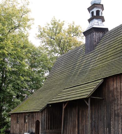 Zdjęcie nr 30: Kościół pomocniczny pw. Wniebowzięcia Najświętszej Marii Panny w Woli Radziszowskiej to niewielkich rozmiarów, orientowana, jednonawowa budowla drewniana, konstrukcji zrębowej, z wieżą w słup. Prezbiterium jest zamknięte wielobocznie, nieco wyższa i szersza nawa ma rzut prostokąta zbliżonego do kwadratu. Do prezbiterium od strony północnej przylega zakrystia, od wschodniej Ogrójec, przy nawie od południa umieszczona została niewielka, prostokątna kruchta. Od zachodu do budowli dostawiona jest czworoboczna wieża o pochyłych ścianach, z podwieszoną izbicą z czworobocznymi występami na przekątnikach; hełm ostrosłupowy z czterema wieżyczkami na występach. W przyziemiu wieży znajduje się kruchta, a dookoła wieży szalowane soboty. Wewnątrz świątyni stropy; w nawie z zaskrzynieniami. Otwór tęczowy ma kształt ostrołuku, belka jest profilowana, na niej umieszczony jest krucyfiks o charakterze późnogotyckim. Chór muzyczny w zachodniej części nawy jest drewniany, wsparty na dwóch kolumnach. Okna nawy i prezbiterium są głównie prostokątne zamknięte półkoliście; z witrażami (z wyjątkiem okna za ołtarzem głównym); ponadto budowlę doświetlają niewielkie okienka prostokątne. Do świątyni prowadzi dwoje drzwi ostrołukowych, trzecie od strony zachodniej przerobiono w 1933 roku. Dachy kościoła są kryte gontem, ponad nawą i kruchtą południową dwuspadowe, nad zakrystią jednospadowy, nad prezbiterium wielopołaciowy. Wieżyczka na sygnaturkę o formach barokowych z glorietą, cebulastym hełmem zwieńczonym kulą oraz krzyżem; kryta blachą. Świątynia usytuowana jest na niewielkiej działce, wydzielonej ogrodzeniem z drewnianych bali, krytym gontem. Bramy wiodące na posesję umieszczono od zachodu oraz w południowo-wschodnim narożniku działki.