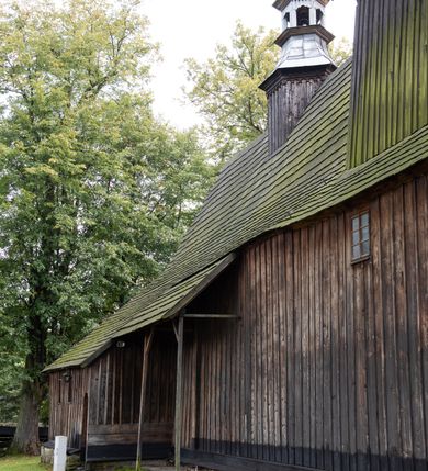 Zdjęcie nr 34: Kościół pomocniczny pw. Wniebowzięcia Najświętszej Marii Panny w Woli Radziszowskiej to niewielkich rozmiarów, orientowana, jednonawowa budowla drewniana, konstrukcji zrębowej, z wieżą w słup. Prezbiterium jest zamknięte wielobocznie, nieco wyższa i szersza nawa ma rzut prostokąta zbliżonego do kwadratu. Do prezbiterium od strony północnej przylega zakrystia, od wschodniej Ogrójec, przy nawie od południa umieszczona została niewielka, prostokątna kruchta. Od zachodu do budowli dostawiona jest czworoboczna wieża o pochyłych ścianach, z podwieszoną izbicą z czworobocznymi występami na przekątnikach; hełm ostrosłupowy z czterema wieżyczkami na występach. W przyziemiu wieży znajduje się kruchta, a dookoła wieży szalowane soboty. Wewnątrz świątyni stropy; w nawie z zaskrzynieniami. Otwór tęczowy ma kształt ostrołuku, belka jest profilowana, na niej umieszczony jest krucyfiks o charakterze późnogotyckim. Chór muzyczny w zachodniej części nawy jest drewniany, wsparty na dwóch kolumnach. Okna nawy i prezbiterium są głównie prostokątne zamknięte półkoliście; z witrażami (z wyjątkiem okna za ołtarzem głównym); ponadto budowlę doświetlają niewielkie okienka prostokątne. Do świątyni prowadzi dwoje drzwi ostrołukowych, trzecie od strony zachodniej przerobiono w 1933 roku. Dachy kościoła są kryte gontem, ponad nawą i kruchtą południową dwuspadowe, nad zakrystią jednospadowy, nad prezbiterium wielopołaciowy. Wieżyczka na sygnaturkę o formach barokowych z glorietą, cebulastym hełmem zwieńczonym kulą oraz krzyżem; kryta blachą. Świątynia usytuowana jest na niewielkiej działce, wydzielonej ogrodzeniem z drewnianych bali, krytym gontem. Bramy wiodące na posesję umieszczono od zachodu oraz w południowo-wschodnim narożniku działki.