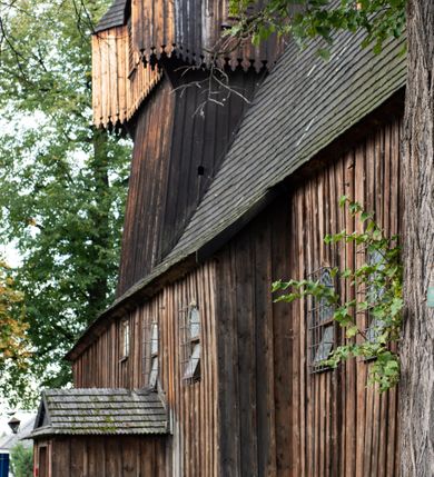 Zdjęcie nr 39: Kościół pomocniczny pw. Wniebowzięcia Najświętszej Marii Panny w Woli Radziszowskiej to niewielkich rozmiarów, orientowana, jednonawowa budowla drewniana, konstrukcji zrębowej, z wieżą w słup. Prezbiterium jest zamknięte wielobocznie, nieco wyższa i szersza nawa ma rzut prostokąta zbliżonego do kwadratu. Do prezbiterium od strony północnej przylega zakrystia, od wschodniej Ogrójec, przy nawie od południa umieszczona została niewielka, prostokątna kruchta. Od zachodu do budowli dostawiona jest czworoboczna wieża o pochyłych ścianach, z podwieszoną izbicą z czworobocznymi występami na przekątnikach; hełm ostrosłupowy z czterema wieżyczkami na występach. W przyziemiu wieży znajduje się kruchta, a dookoła wieży szalowane soboty. Wewnątrz świątyni stropy; w nawie z zaskrzynieniami. Otwór tęczowy ma kształt ostrołuku, belka jest profilowana, na niej umieszczony jest krucyfiks o charakterze późnogotyckim. Chór muzyczny w zachodniej części nawy jest drewniany, wsparty na dwóch kolumnach. Okna nawy i prezbiterium są głównie prostokątne zamknięte półkoliście; z witrażami (z wyjątkiem okna za ołtarzem głównym); ponadto budowlę doświetlają niewielkie okienka prostokątne. Do świątyni prowadzi dwoje drzwi ostrołukowych, trzecie od strony zachodniej przerobiono w 1933 roku. Dachy kościoła są kryte gontem, ponad nawą i kruchtą południową dwuspadowe, nad zakrystią jednospadowy, nad prezbiterium wielopołaciowy. Wieżyczka na sygnaturkę o formach barokowych z glorietą, cebulastym hełmem zwieńczonym kulą oraz krzyżem; kryta blachą. Świątynia usytuowana jest na niewielkiej działce, wydzielonej ogrodzeniem z drewnianych bali, krytym gontem. Bramy wiodące na posesję umieszczono od zachodu oraz w południowo-wschodnim narożniku działki.