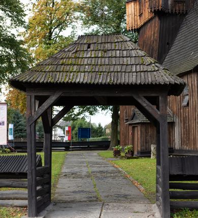 Zdjęcie nr 40: Kościół pomocniczny pw. Wniebowzięcia Najświętszej Marii Panny w Woli Radziszowskiej to niewielkich rozmiarów, orientowana, jednonawowa budowla drewniana, konstrukcji zrębowej, z wieżą w słup. Prezbiterium jest zamknięte wielobocznie, nieco wyższa i szersza nawa ma rzut prostokąta zbliżonego do kwadratu. Do prezbiterium od strony północnej przylega zakrystia, od wschodniej Ogrójec, przy nawie od południa umieszczona została niewielka, prostokątna kruchta. Od zachodu do budowli dostawiona jest czworoboczna wieża o pochyłych ścianach, z podwieszoną izbicą z czworobocznymi występami na przekątnikach; hełm ostrosłupowy z czterema wieżyczkami na występach. W przyziemiu wieży znajduje się kruchta, a dookoła wieży szalowane soboty. Wewnątrz świątyni stropy; w nawie z zaskrzynieniami. Otwór tęczowy ma kształt ostrołuku, belka jest profilowana, na niej umieszczony jest krucyfiks o charakterze późnogotyckim. Chór muzyczny w zachodniej części nawy jest drewniany, wsparty na dwóch kolumnach. Okna nawy i prezbiterium są głównie prostokątne zamknięte półkoliście; z witrażami (z wyjątkiem okna za ołtarzem głównym); ponadto budowlę doświetlają niewielkie okienka prostokątne. Do świątyni prowadzi dwoje drzwi ostrołukowych, trzecie od strony zachodniej przerobiono w 1933 roku. Dachy kościoła są kryte gontem, ponad nawą i kruchtą południową dwuspadowe, nad zakrystią jednospadowy, nad prezbiterium wielopołaciowy. Wieżyczka na sygnaturkę o formach barokowych z glorietą, cebulastym hełmem zwieńczonym kulą oraz krzyżem; kryta blachą. Świątynia usytuowana jest na niewielkiej działce, wydzielonej ogrodzeniem z drewnianych bali, krytym gontem. Bramy wiodące na posesję umieszczono od zachodu oraz w południowo-wschodnim narożniku działki.