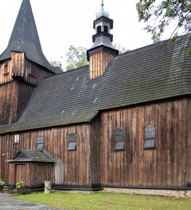 Zdjęcie nr 43: Kościół pomocniczny pw. Wniebowzięcia Najświętszej Marii Panny w Woli Radziszowskiej to niewielkich rozmiarów, orientowana, jednonawowa budowla drewniana, konstrukcji zrębowej, z wieżą w słup. Prezbiterium jest zamknięte wielobocznie, nieco wyższa i szersza nawa ma rzut prostokąta zbliżonego do kwadratu. Do prezbiterium od strony północnej przylega zakrystia, od wschodniej Ogrójec, przy nawie od południa umieszczona została niewielka, prostokątna kruchta. Od zachodu do budowli dostawiona jest czworoboczna wieża o pochyłych ścianach, z podwieszoną izbicą z czworobocznymi występami na przekątnikach; hełm ostrosłupowy z czterema wieżyczkami na występach. W przyziemiu wieży znajduje się kruchta, a dookoła wieży szalowane soboty. Wewnątrz świątyni stropy; w nawie z zaskrzynieniami. Otwór tęczowy ma kształt ostrołuku, belka jest profilowana, na niej umieszczony jest krucyfiks o charakterze późnogotyckim. Chór muzyczny w zachodniej części nawy jest drewniany, wsparty na dwóch kolumnach. Okna nawy i prezbiterium są głównie prostokątne zamknięte półkoliście; z witrażami (z wyjątkiem okna za ołtarzem głównym); ponadto budowlę doświetlają niewielkie okienka prostokątne. Do świątyni prowadzi dwoje drzwi ostrołukowych, trzecie od strony zachodniej przerobiono w 1933 roku. Dachy kościoła są kryte gontem, ponad nawą i kruchtą południową dwuspadowe, nad zakrystią jednospadowy, nad prezbiterium wielopołaciowy. Wieżyczka na sygnaturkę o formach barokowych z glorietą, cebulastym hełmem zwieńczonym kulą oraz krzyżem; kryta blachą. Świątynia usytuowana jest na niewielkiej działce, wydzielonej ogrodzeniem z drewnianych bali, krytym gontem. Bramy wiodące na posesję umieszczono od zachodu oraz w południowo-wschodnim narożniku działki.