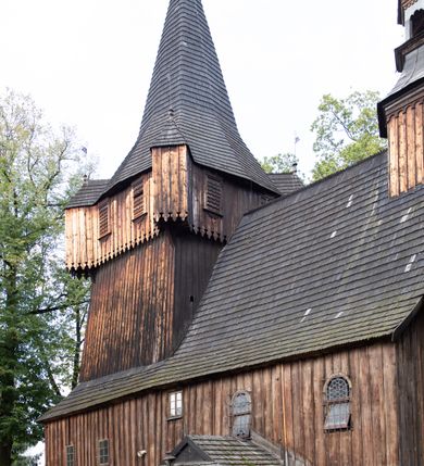 Zdjęcie nr 42: Kościół pomocniczny pw. Wniebowzięcia Najświętszej Marii Panny w Woli Radziszowskiej to niewielkich rozmiarów, orientowana, jednonawowa budowla drewniana, konstrukcji zrębowej, z wieżą w słup. Prezbiterium jest zamknięte wielobocznie, nieco wyższa i szersza nawa ma rzut prostokąta zbliżonego do kwadratu. Do prezbiterium od strony północnej przylega zakrystia, od wschodniej Ogrójec, przy nawie od południa umieszczona została niewielka, prostokątna kruchta. Od zachodu do budowli dostawiona jest czworoboczna wieża o pochyłych ścianach, z podwieszoną izbicą z czworobocznymi występami na przekątnikach; hełm ostrosłupowy z czterema wieżyczkami na występach. W przyziemiu wieży znajduje się kruchta, a dookoła wieży szalowane soboty. Wewnątrz świątyni stropy; w nawie z zaskrzynieniami. Otwór tęczowy ma kształt ostrołuku, belka jest profilowana, na niej umieszczony jest krucyfiks o charakterze późnogotyckim. Chór muzyczny w zachodniej części nawy jest drewniany, wsparty na dwóch kolumnach. Okna nawy i prezbiterium są głównie prostokątne zamknięte półkoliście; z witrażami (z wyjątkiem okna za ołtarzem głównym); ponadto budowlę doświetlają niewielkie okienka prostokątne. Do świątyni prowadzi dwoje drzwi ostrołukowych, trzecie od strony zachodniej przerobiono w 1933 roku. Dachy kościoła są kryte gontem, ponad nawą i kruchtą południową dwuspadowe, nad zakrystią jednospadowy, nad prezbiterium wielopołaciowy. Wieżyczka na sygnaturkę o formach barokowych z glorietą, cebulastym hełmem zwieńczonym kulą oraz krzyżem; kryta blachą. Świątynia usytuowana jest na niewielkiej działce, wydzielonej ogrodzeniem z drewnianych bali, krytym gontem. Bramy wiodące na posesję umieszczono od zachodu oraz w południowo-wschodnim narożniku działki.