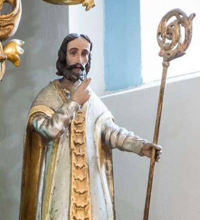 Zdjęcie nr 1: Figura niemal pełnoplastyczna, z tyłu ścięta i drążona ukazuje świętego, przedstawionego w pozycji stojącej. Święty w lewej dłoni trzyma oparty o ziemię pastorał, a prawą wznosi ugiętą. Ma pociągłą twarz, duże oczy, nos długi o prostym profilu i lekko rozchylone usta. Twarz ma okoloną brązowymi włosami formowanymi w pukle i krótką brodą. Jest ubrany w srebrną albę i biały ornat ze złotą pretekstą dekorowaną cekinami i złotym lamowaniem. Polichromia w partiach ciała naturalistyczna.