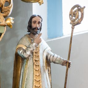 Zdjęcie nr 1: Figura niemal pełnoplastyczna, z tyłu ścięta i drążona ukazuje świętego, przedstawionego w pozycji stojącej. Święty w lewej dłoni trzyma oparty o ziemię pastorał, a prawą wznosi ugiętą. Ma pociągłą twarz, duże oczy, nos długi o prostym profilu i lekko rozchylone usta. Twarz ma okoloną brązowymi włosami formowanymi w pukle i krótką brodą. Jest ubrany w srebrną albę i biały ornat ze złotą pretekstą dekorowaną cekinami i złotym lamowaniem. Polichromia w partiach ciała naturalistyczna.