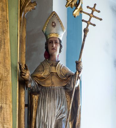 Zdjęcie nr 1: Figura niemal pełnoplastyczna, z tyłu ścięta i drążona. Święty Stanisław został ukazany w pozycji stojącej z uniesionymi rękami, w lewej dłoni trzymając krzyż patriarszy. Twarz ma pociągłą, o drobnych rysach. Jest ubrany w jasnoróżową sutannę, srebrną komżę oraz złote mucet i kapę. Na głowie ma infułę, na dłoniach rękawiczki. Po lewej stronie, u stóp biskupa ukazany został Piotrowin, w popiersiu, z głową wzniesioną w stronę św. Stanisława, z rękami złożonymi w geście modlitewnym. Polichromia w odsłoniętych partiach ciała naturalistyczna. 