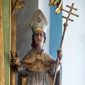 Zdjęcie nr 1: Figura niemal pełnoplastyczna, z tyłu ścięta i drążona. Święty Stanisław został ukazany w pozycji stojącej z uniesionymi rękami, w lewej dłoni trzymając krzyż patriarszy. Twarz ma pociągłą, o drobnych rysach. Jest ubrany w jasnoróżową sutannę, srebrną komżę oraz złote mucet i kapę. Na głowie ma infułę, na dłoniach rękawiczki. Po lewej stronie, u stóp biskupa ukazany został Piotrowin, w popiersiu, z głową wzniesioną w stronę św. Stanisława, z rękami złożonymi w geście modlitewnym. Polichromia w odsłoniętych partiach ciała naturalistyczna. 