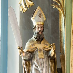 Zdjęcie nr 1: Figura niemal pełnoplastyczna, z tyłu ścięta i drążona. Święty Wojciech został ukazany w pozycji stojącej z uniesionymi rękami, w prawej dłoni trzymając wiosło. Twarz ma pociągłą, brwi uniesione, oczy szeroko otwarte, nos prosty, brodę długą, dzielącą się w dwa pukle. Jest ubrany w jasnoróżową sutannę, srebrną komżę oraz złotą kapę i paliusz. Na głowie ma infułę, na dłoniach rękawiczki. Polichromia w partiach ciała naturalistyczna. 