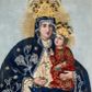 Zdjęcie nr 1: Obraz w kształcie stojącego prostokąta z przedstawieniem Matki Boskiej z Dzieciątkiem. Twarze, dłonie i tło są malowane, szaty i korony, wykonane z dekorowanego haftem i kamieniami materiału, zostały nałożone na obraz. Maria spogląda na widza, na lewej ręce trzyma Dzieciątko, w prawej ma różaniec, jej głowa jest nieproporcjonalnie mała w stosunku do okrytego szatami ciała. Ma owalną, delikatną twarz o szeroko otwartych ciemnych oczach, wąskim, długim nosie i małych ustach. Jest ubrana w kremową suknię marszczoną pod szyją i na rękawach, związaną na dekolcie tasiemką na kokardkę ozdobioną zielonym kamieniem.  Na suknię ma nałożony granatowy, zarzucony na głowę płaszcz lamowany ornamentem z wici utworzonej ze sznurka i koralików, z których wykonano również dekorujące poły kwiaty i gwiazdki. Dzieciątko siedzi na jej ręce, patrzy przed siebie, prawą ręką czyni gest błogosławieństwa, w lewej trzyma książeczkę. Ma podłużną twarz, duże oczy, wąski nos i małe usta. Okalają ją ciemne loki.  Jezus jest ubrany w czerwoną suknię z wykonaną ze sznurka i koralików dekoracją kwiatową. Obie postaci mają na głowach korony zamknięte skomponowane z nakładanych sznurków, korali i kamieni, w centrum korony Marii jej monogram, w centrum korony Chrystusa „IHS”. Za głowami malowane nimby promieniste, tło jednolite – szare. 