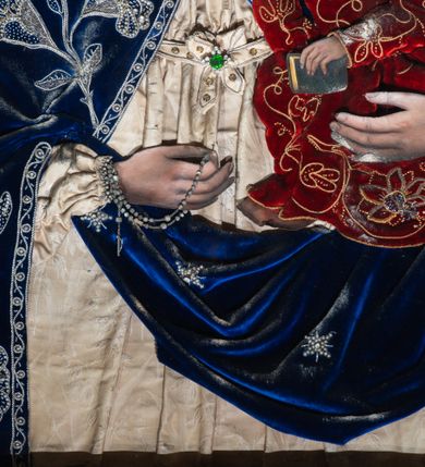 Zdjęcie nr 3: Obraz w kształcie stojącego prostokąta z przedstawieniem Matki Boskiej z Dzieciątkiem. Twarze, dłonie i tło są malowane, szaty i korony, wykonane z dekorowanego haftem i kamieniami materiału, zostały nałożone na obraz. Maria spogląda na widza, na lewej ręce trzyma Dzieciątko, w prawej ma różaniec, jej głowa jest nieproporcjonalnie mała w stosunku do okrytego szatami ciała. Ma owalną, delikatną twarz o szeroko otwartych ciemnych oczach, wąskim, długim nosie i małych ustach. Jest ubrana w kremową suknię marszczoną pod szyją i na rękawach, związaną na dekolcie tasiemką na kokardkę ozdobioną zielonym kamieniem.  Na suknię ma nałożony granatowy, zarzucony na głowę płaszcz lamowany ornamentem z wici utworzonej ze sznurka i koralików, z których wykonano również dekorujące poły kwiaty i gwiazdki. Dzieciątko siedzi na jej ręce, patrzy przed siebie, prawą ręką czyni gest błogosławieństwa, w lewej trzyma książeczkę. Ma podłużną twarz, duże oczy, wąski nos i małe usta. Okalają ją ciemne loki.  Jezus jest ubrany w czerwoną suknię z wykonaną ze sznurka i koralików dekoracją kwiatową. Obie postaci mają na głowach korony zamknięte skomponowane z nakładanych sznurków, korali i kamieni, w centrum korony Marii jej monogram, w centrum korony Chrystusa „IHS”. Za głowami malowane nimby promieniste, tło jednolite – szare. 