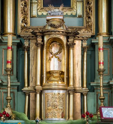 Zdjęcie nr 1: Tabernakulum o rozbudowanej, dwustrefowej strukturze, na planie wklęsło-wypukłym. Dolna kondygnacja niższa, z wypukłymi drzwiczkami pośrodku, górna z ulokowanym nad nimi tronem wystawienia w formie wnęki zamkniętej łukiem półkolistym. Każdą  kondygnację wieńczy belkowanie – w dolnej partii fryz i gzyms, w górnej pełne z impostami i silnie wysuniętym gzymsem – wyłamane na osiach ułożonych skośnie, smukłych kolumienek korynckich. W zwieńczeniu niewielka kopuła, na której ustawiona jest rzeźba baranka spoczywającego na zamkniętej księdze, na tle obłoków i glorii. Na drzwiczkach tabernakulum znajduje się płaskorzeźbione przedstawienie Chrystusa siedzącego za stołem, przed chlebem i kielichem, między ustawionymi symetrycznie świecznikami. W niszę powyżej wstawiono postument dekorowany podwieszoną wstęgą oraz wieniec z owoców i kwiatów przewiązany wstęgami o karbowanych końcach opadających ku dołowi. Na fragmentach architrawu palmety, na kopule girlanda z pąków róż. Całość struktury złocona i srebrzona.